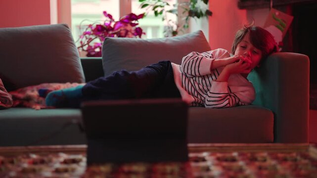 Tired young boy resting on sofa and watching tablet after long day, peaceful home scene highlighting digital entertainment and comfort