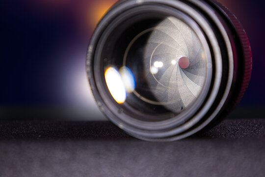 Detailed close-up image showing the camera lens with visible aperture blades and reflections on the glass surface. Focus on the internal structure and curvature of the lens elements, highlighting the