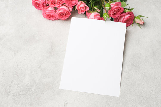 Blank paper card mockup placed on a textured gray surface with pink roses arranged beside it, ideal for design presentations or greeting card concepts
