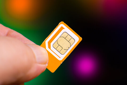 Close-up of a hand holding an orange SIM card against a vibrant, blurred backdrop featuring green, purple, and orange bokeh lights. Sharp focus on the SIM cards gold chip and detailed circuitry, highl