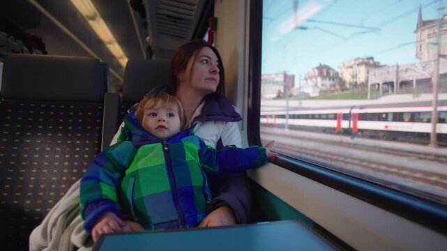 Mother holding toddler beside train window during rail journey, child looking toward camera while parent watches passing countryside in quiet travel moment