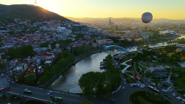 Golden hour aerial view of Tbilisi, Georgia, featuring the Kura River, the Bridge of Peace, and a hot air balloon over Rike Park with the city skyline at sunset