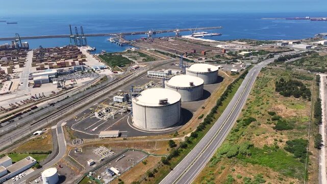 Aerial view of lng terminal with multiple gas storage tanks. Nearby industrial area includes ships and energy infrastructure. Focus on natural gas supply and storage.