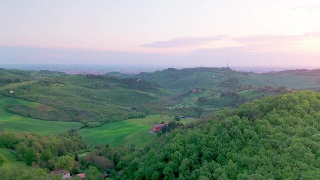 Drone rising at dawn over Parco di Forte Bandiera on Monte Donato, revealing green hills, forest, and distant Bologna as the camera rotates in a half‑circle.