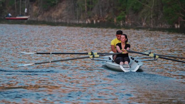 Close cinematic view of two rowers gliding across a calm lake surrounded by rocky hills and pine trees during sunset. Peaceful outdoor sports scene with teamwork and motion on freshwater.