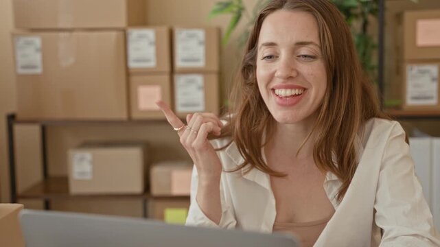 Woman points finger at a parcel and smiles at a laptop, surrounded by stacked boxes and shelving in a building office; small business joy.