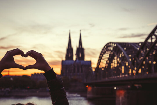Germany, Cologne, woman shaping heart with her hands in front of Cologne Cathedral and Hohenzollern Bridge at dusk
