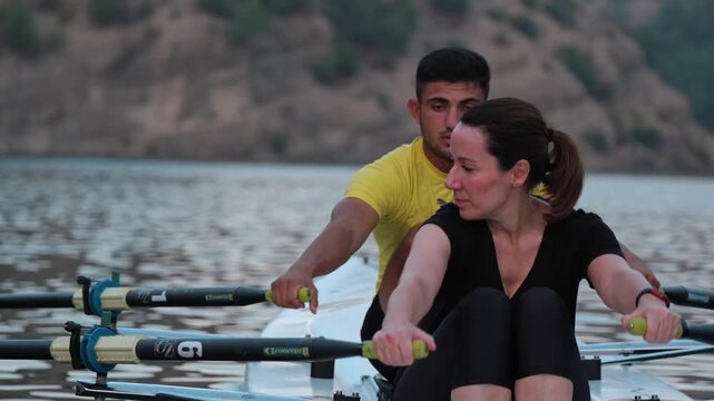 Close cinematic view of two rowers gliding across a calm lake surrounded by rocky hills and pine trees during sunset. Peaceful outdoor sports scene with teamwork and motion on freshwater.