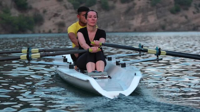 Close cinematic view of two rowers gliding across a calm lake surrounded by rocky hills and pine trees during sunset. Peaceful outdoor sports scene with teamwork and motion on freshwater.