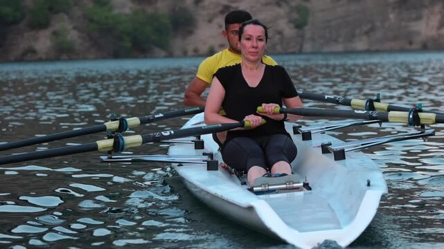 Close cinematic view of two rowers gliding across a calm lake surrounded by rocky hills and pine trees during sunset. Peaceful outdoor sports scene with teamwork and motion on freshwater.