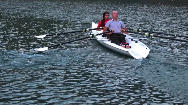 Close cinematic view of two rowers gliding across a calm lake surrounded by rocky hills and pine trees during sunset. Peaceful outdoor sports scene with teamwork and motion on freshwater.
