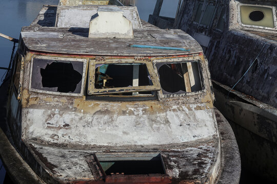Abandoned weathered fishing boat with broken cabin windows and deteriorated wooden surfaces beside a quiet waterfront.