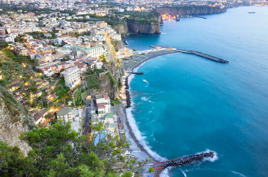 Aerial sunset view of Meta di Sorrento coastal town, Italy, overlooking turquoise Mediterranean waters. Shoreline of Sorrento Peninsula with colorful buildings and protective breakwaters along bay