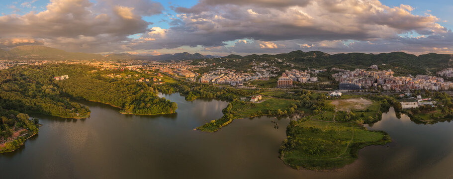 Panoramic drone photo of Tirana Lake, Albania, showing calm water, green parkland, city skyline, hills, and dramatic evening clouds.