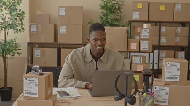 Man cheering with raised fists over laptop and parcels in a building packing area; small business joy.