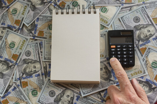 A hand pressing the buttons of a calculator next to a blank paper notebook against a background of U.S. hundred-dollar bills, ideal for adding text or a message.