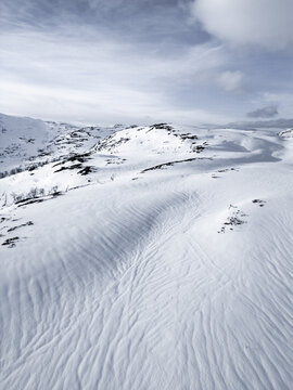 Aerial view of snow-covered mountain slopes with wind-blown textures and rolling hills under a cloudy winter sky in Kvaloya, Troms, Norway.