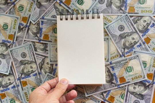 A hand holding a blank paper notebook against a background of U.S. hundred-dollar bills, ideal for adding text or a message.