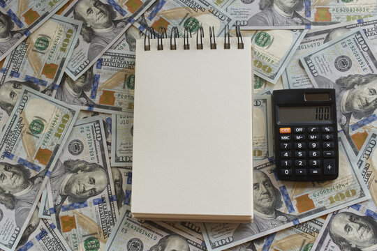 A blank paper notebook and calculator against a background of U.S. hundred-dollar bills, ideal for adding text or a message.