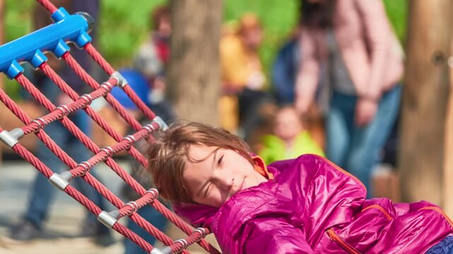 A girl in a red jacket is swinging on a hammock in a city park.
