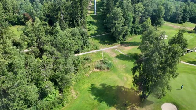 Scenic aerial view of a vibrant green golf course nestled in an alpine forest with a single golfer putting on the green next to a sand bunker in summer