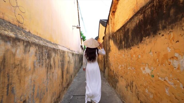 Asian woman in an ao dai costume walks in an alley in Hoi An, ancient town.