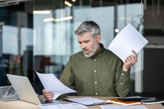 Stressed businessman at cluttered desk, holding documents and gazing at spreadsheets and reports, overwhelmed by paperwork, financial pressures and looming deadlines