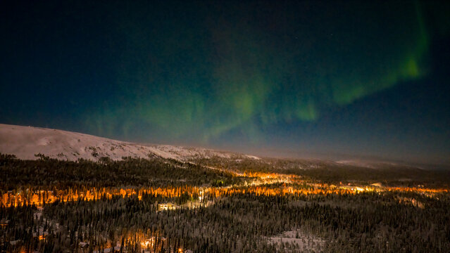 Aerial view of Luosto with the Aurora Borealis dancing over snow-covered forests and the glowing lights of the resort at night Sodankyla, Lapland, Finland.
