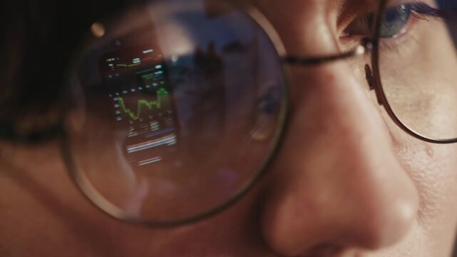 Close up of a woman face wearing glasses with smartphone screen reflected in the lenses. The reflection shows a stock trading app interface. Concept of online trading finance technology investment