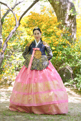 Fototapeta premium Woman in vibrant traditional Korean attire stands gracefully. Her dark jacket is adorned with gold embroidery. The pink skirt features layered, intricate golden patterns