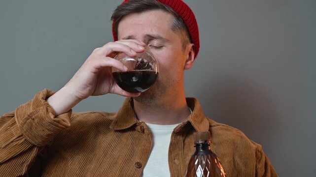 A man in casual attire holds a glass and a bottle, taking a drink in a well-lit indoor setting. He shows different expressions as he tastes the beverage, suggesting a moment of reflection