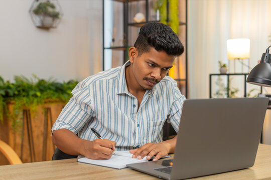 Indian man at home studies online course with laptop and notebook writes notes completes test. Arabian guy at table watches lecture reviews slides uses submits exam checks score and final grade.