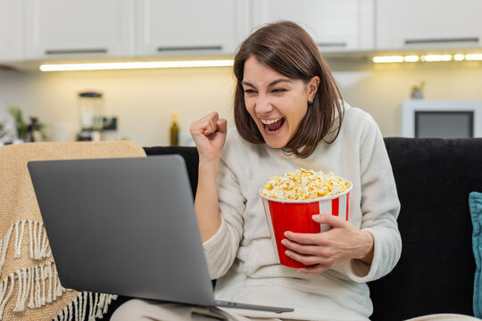 Young woman at home watches football on laptop jumps with joy at goal claps shows like eats popcorn. Caucasian girl on sofa chants for team checks score waves and enjoys stream highlight live in app
