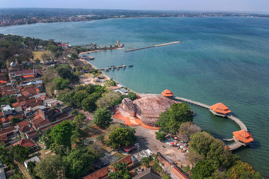Aerial view of the giant turtle-shaped building of Kura-Kura Ocean Park on the coastline with blue sea and urban rooftops in Jepara, Central Java, Indonesia.