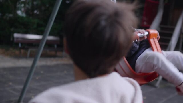 Toddler swinging in bucket seat while older child holds front bar during forward motion at playground showing clear swing frame chains and surrounding park layout