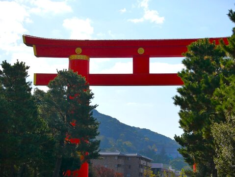 A dramatic low-angle vertical shot of the massive red Torii gate at Heian Jingu Shrine reaching into a beautiful blue sky with soft white clouds.