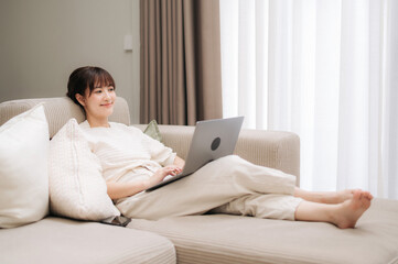Fototapeta premium Beautiful happy young asian woman relaxing on comfortable sofa at home using laptop computer while smiling brightly enjoying peaceful leisure day indoor near bright window wearing casual outfit