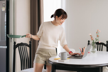 Obraz premium One cheerful young asian woman enjoys multitasking in beautiful home kitchen holding cooking pan and typing on modern laptop while smiling and searching for online recipe