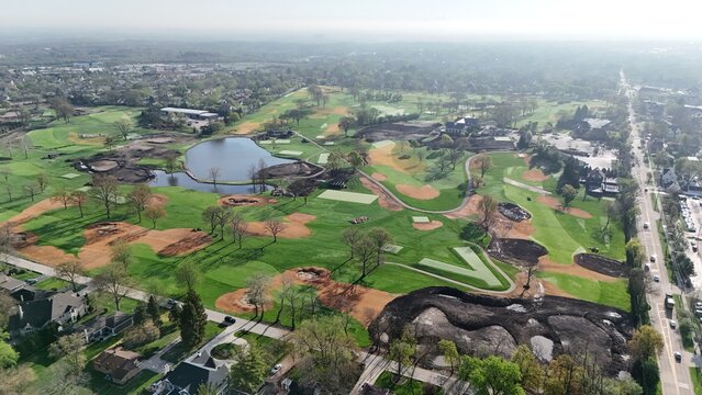Aerial view of a golf course undergoing renovation with green fairways, a blue pond, and patches of dark soil surrounded by suburban houses and a road with cars.