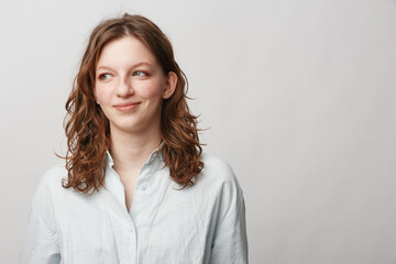 Obraz premium Young woman with curly brown hair wearing white shirt looks sideways with slight smile on light background. Concept of casual style, natural beauty, and positive mood.