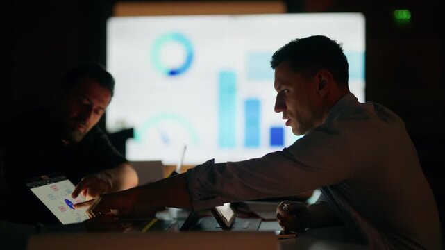 Business people discuss data and strategy in a dark meeting room, utilizing tablets and a large screen with charts. A dedicated team analyzes financial information, working late into the night.