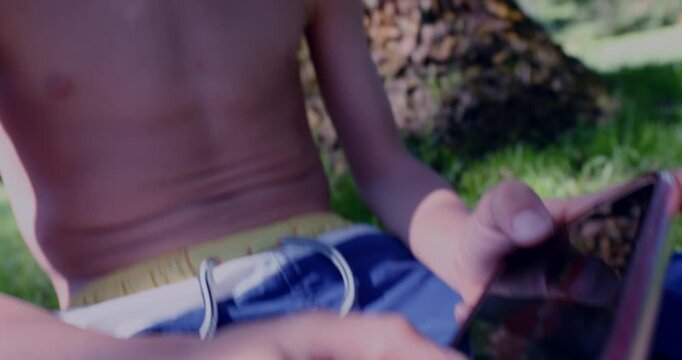 Closeup of shirtless boy&rsquo;s face looking down at smartphone screen while playing game outdoors, framed by tree trunk and soft daylight in lush park setting