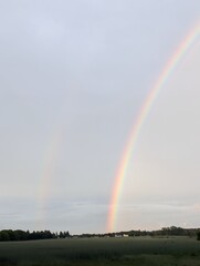 Fototapeta premium A vibrant double rainbow arches across a cloudy sky over a rural landscape with fields and trees below
