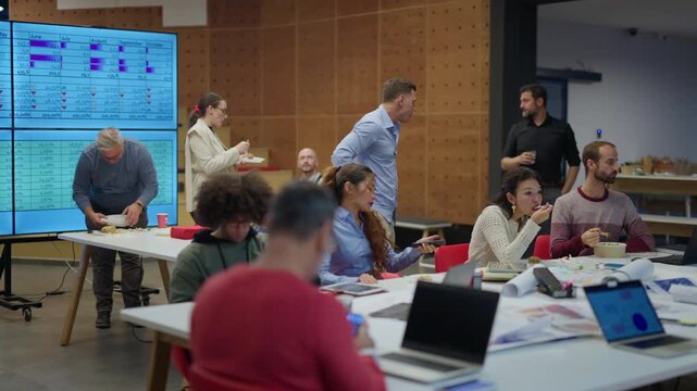 Diverse employees are enjoying a casual lunch break at their modern office workspace. Colleagues gather around a table, eating and socializing during an informal business meeting.