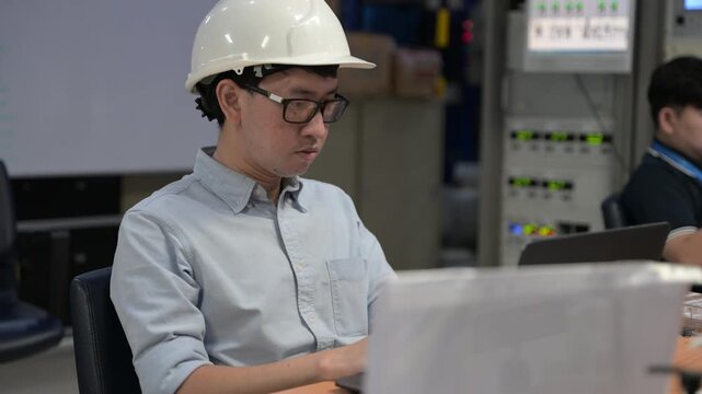 Asian male engineer wearing hardhat coding on laptop in industrial office. Professional technician developer working on software programming for manufacturing automation and factory system.