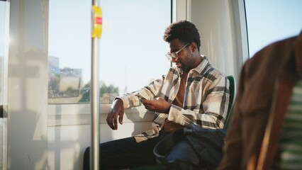 African American man with beard and glasses sitting by window on bus using smartphone. Passenger leaning back relaxed scrolling screen during sunny day ride. Casual moment on public transport. © VAKSMANV