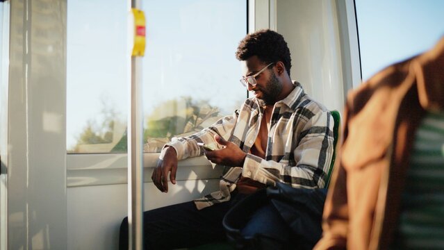 African American man with beard and glasses sitting by window on bus using smartphone. Passenger leaning back relaxed scrolling screen during sunny day ride. Casual moment on public transport.