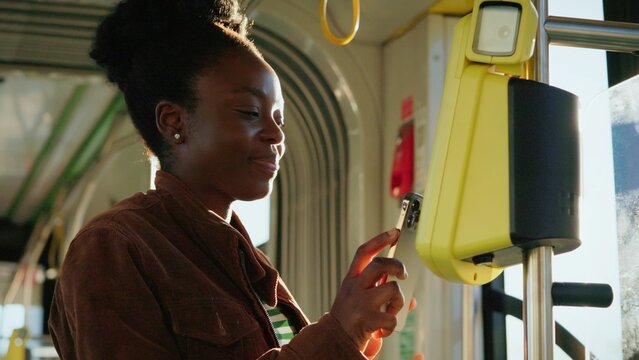 Charming African American woman holding smartphone close to yellow validator paying for ride on bus. Passenger in striped shirt and jacket completing contactless payment. Modern technology.