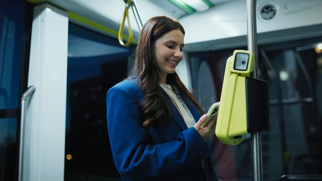 Pretty Caucasian woman with long hair holding smartphone and validating ticket on public transport. Female smiling while using contactless payment system inside modern city bus at night.