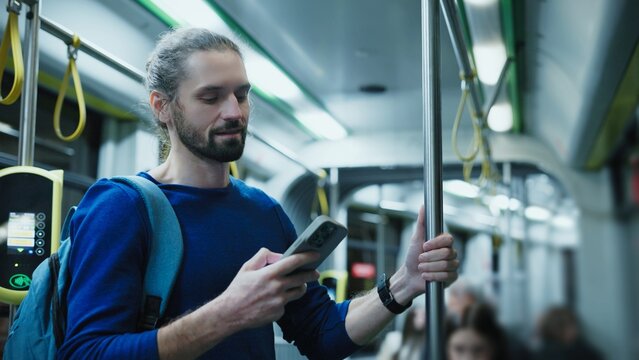Caucasian man with beard wearing blue sweater standing on city bus holding pole with one hand and smartphone in other. Male reading online news during evening commute home from work.